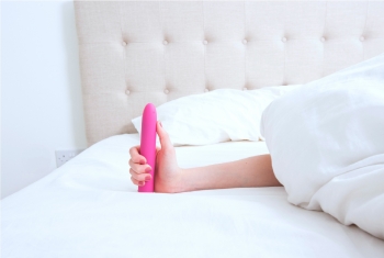 A hand holding a pink vibrator in a white plush bed